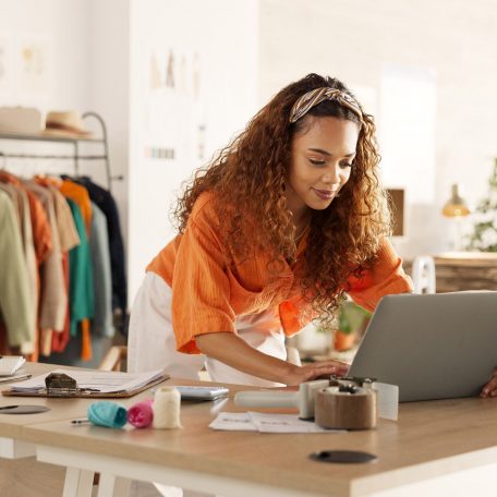 Logistics, laptop and fashion manager of retail or boutique shop checking clothing stock in boxes on a checklist. Small business, entrepreneur and young woman working with clothes inventory in store.